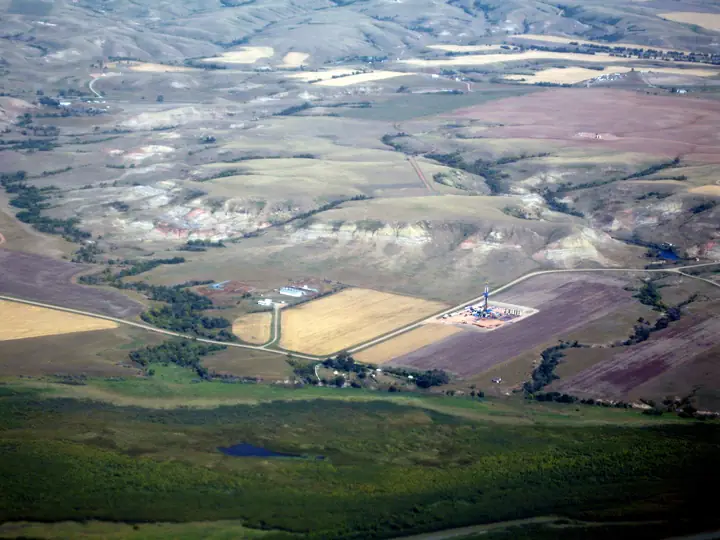 Aerial view of a drilling rig on a small well pad among farm fields and badlands in North Dakota.